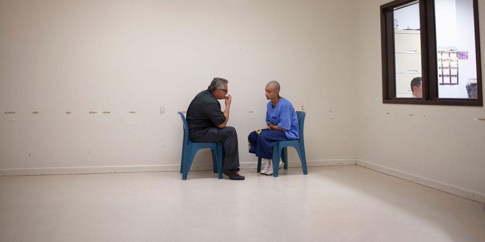 Man and young inmate sitting talking on plastic chairs under fluorescent lights.