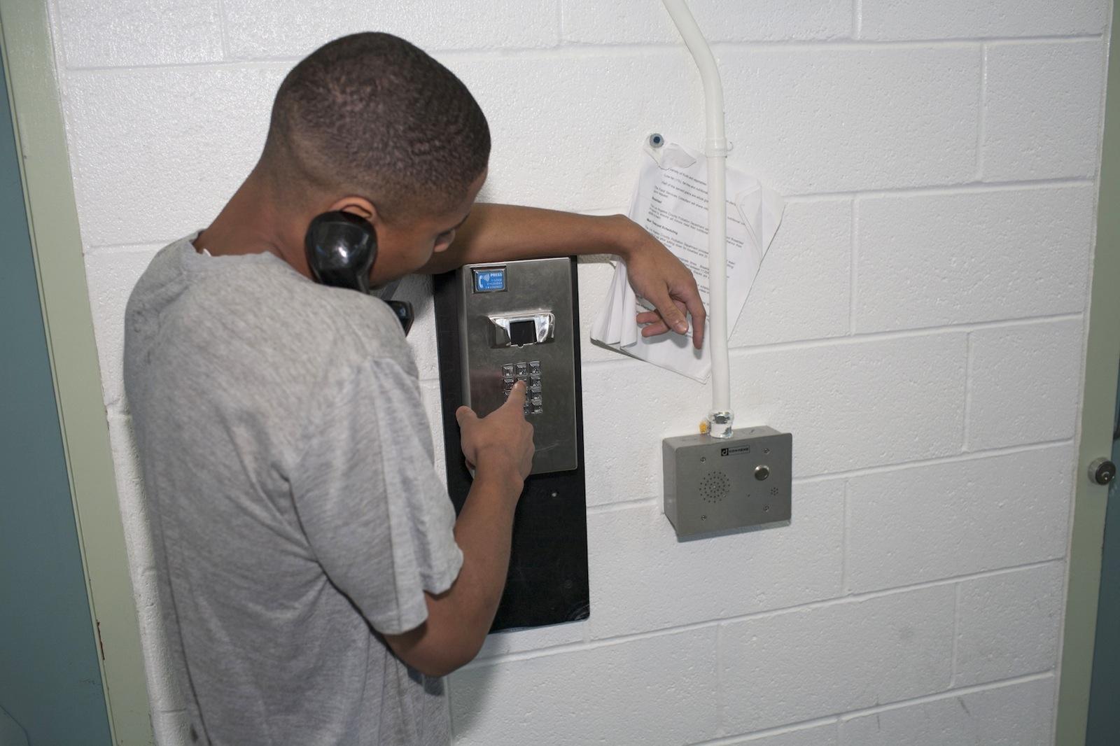 Young black male using a pay phone hanging on a white tiled wall. 