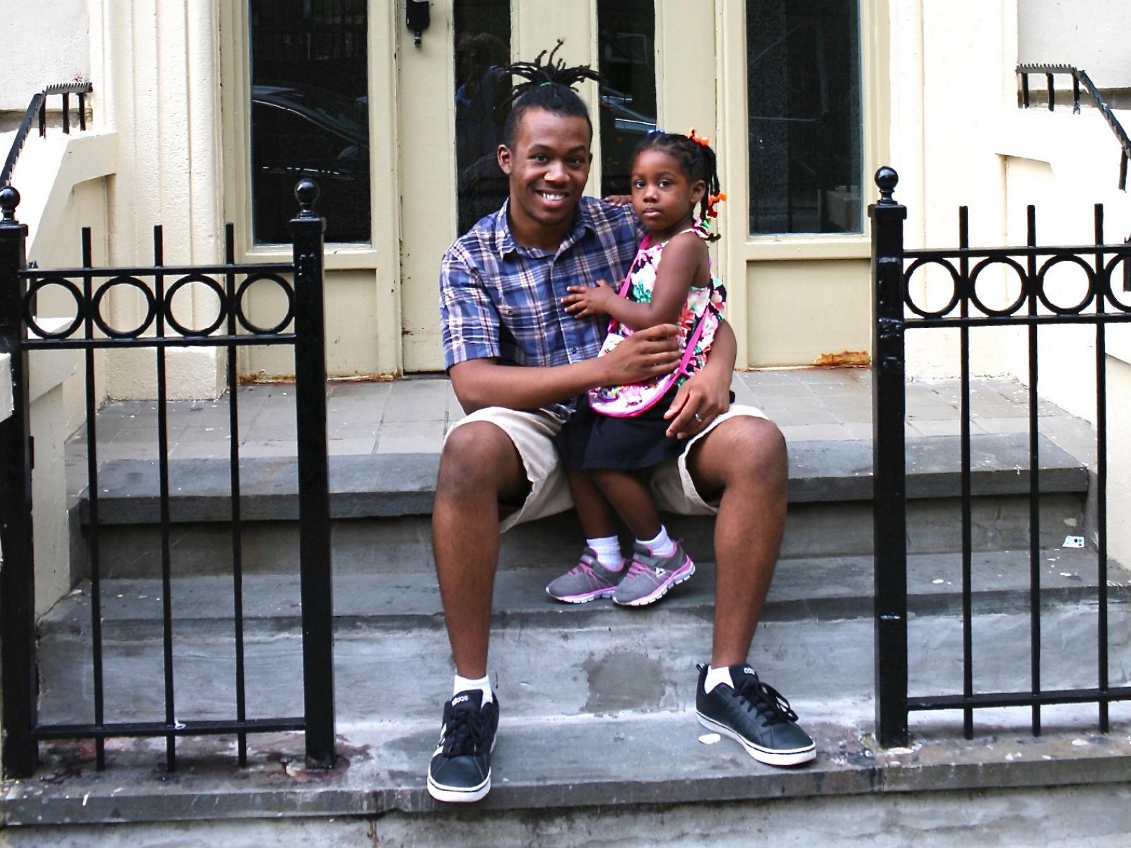 Jordan, a young Black man, sitting outside his house with small girl on his knee. 