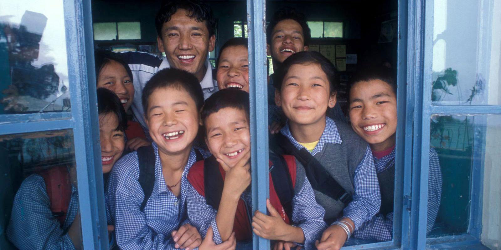 Group of children in school uniforms looking out a window, smiling.
