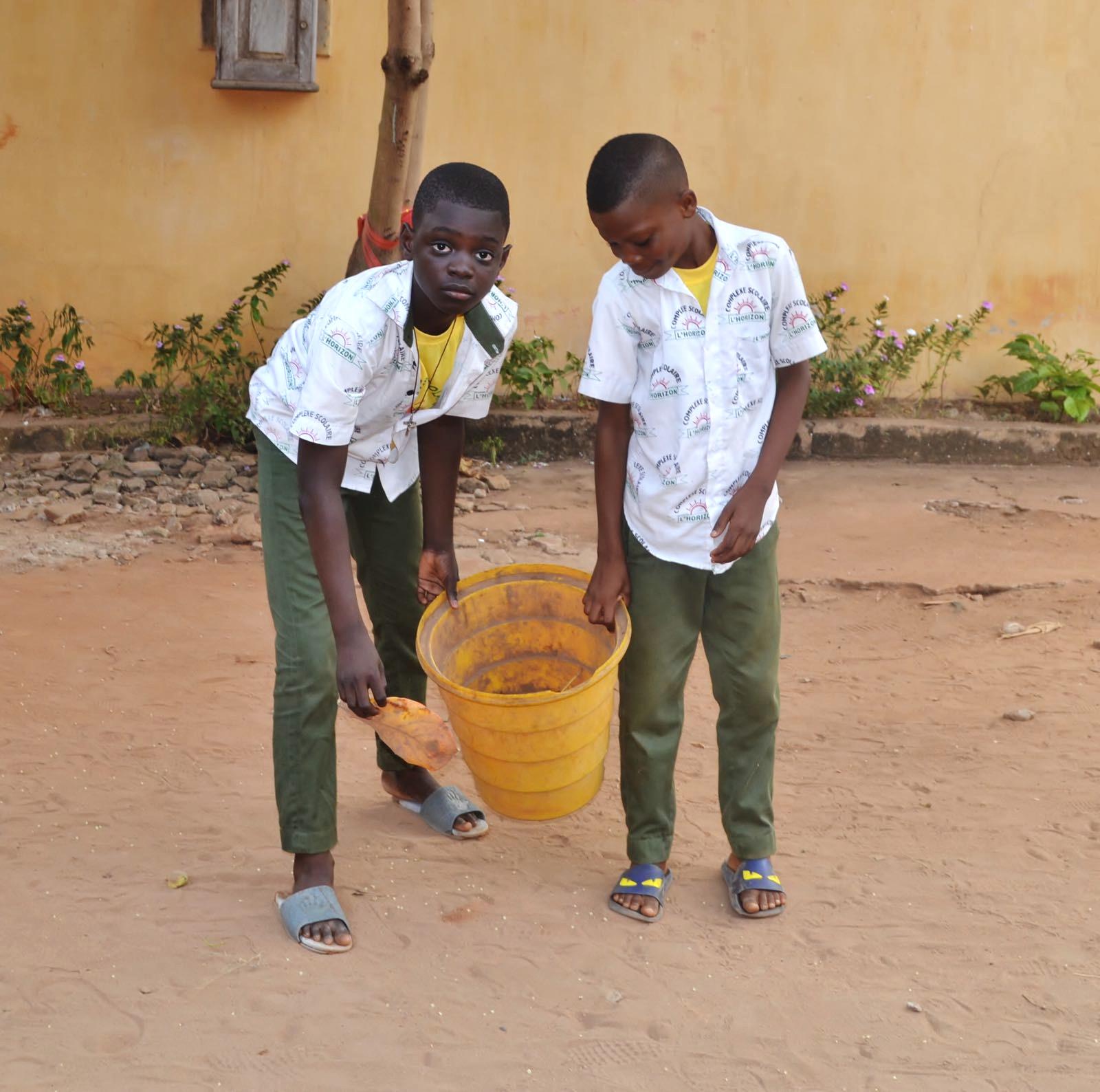 Two boys in green pants and white shirts carrying a bucket