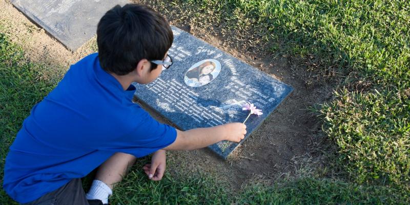 Small boy placing flower on young woman’s grave marker.