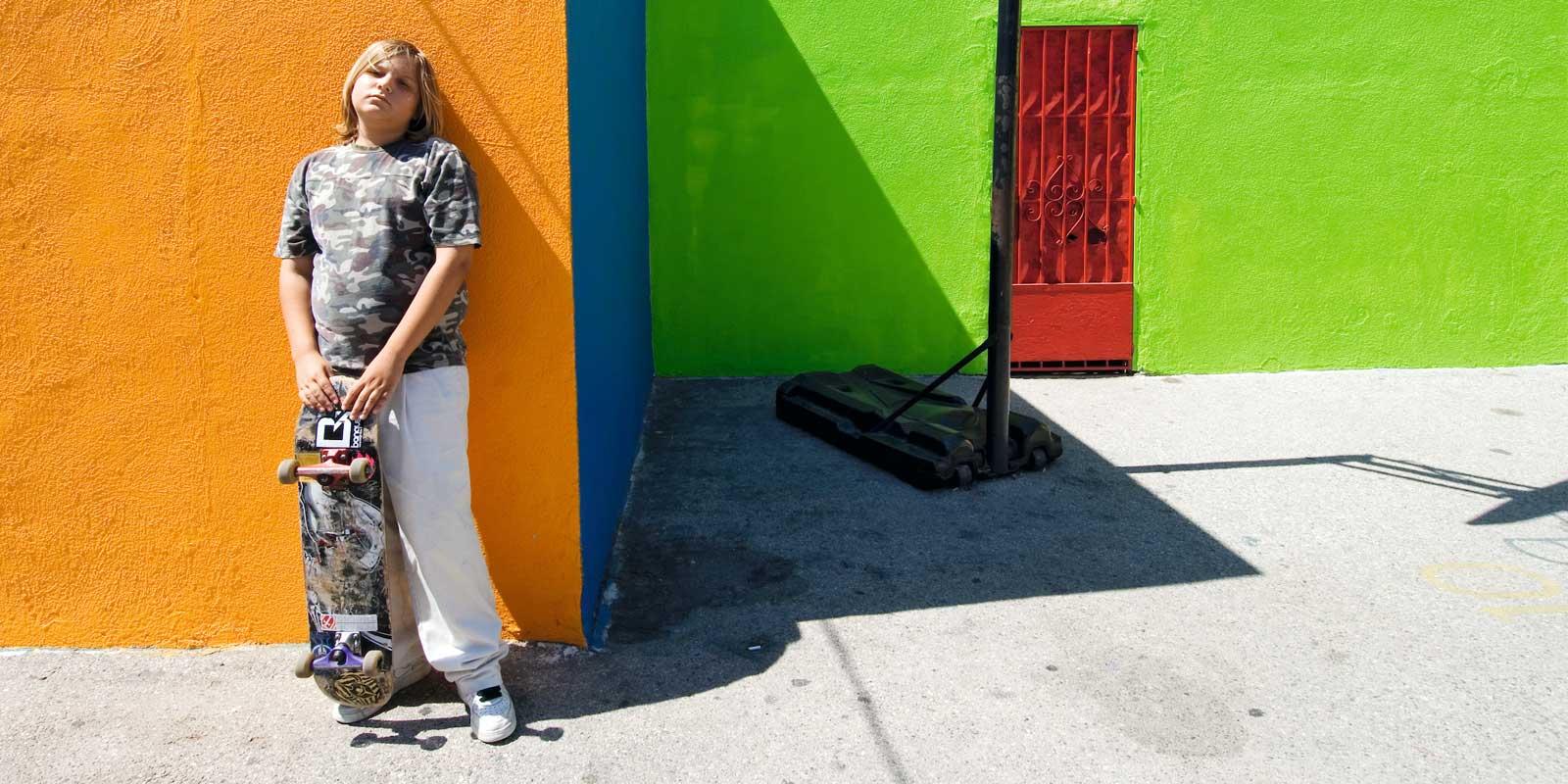 Boy with blond hair and skateboard in front of colorful buildings.