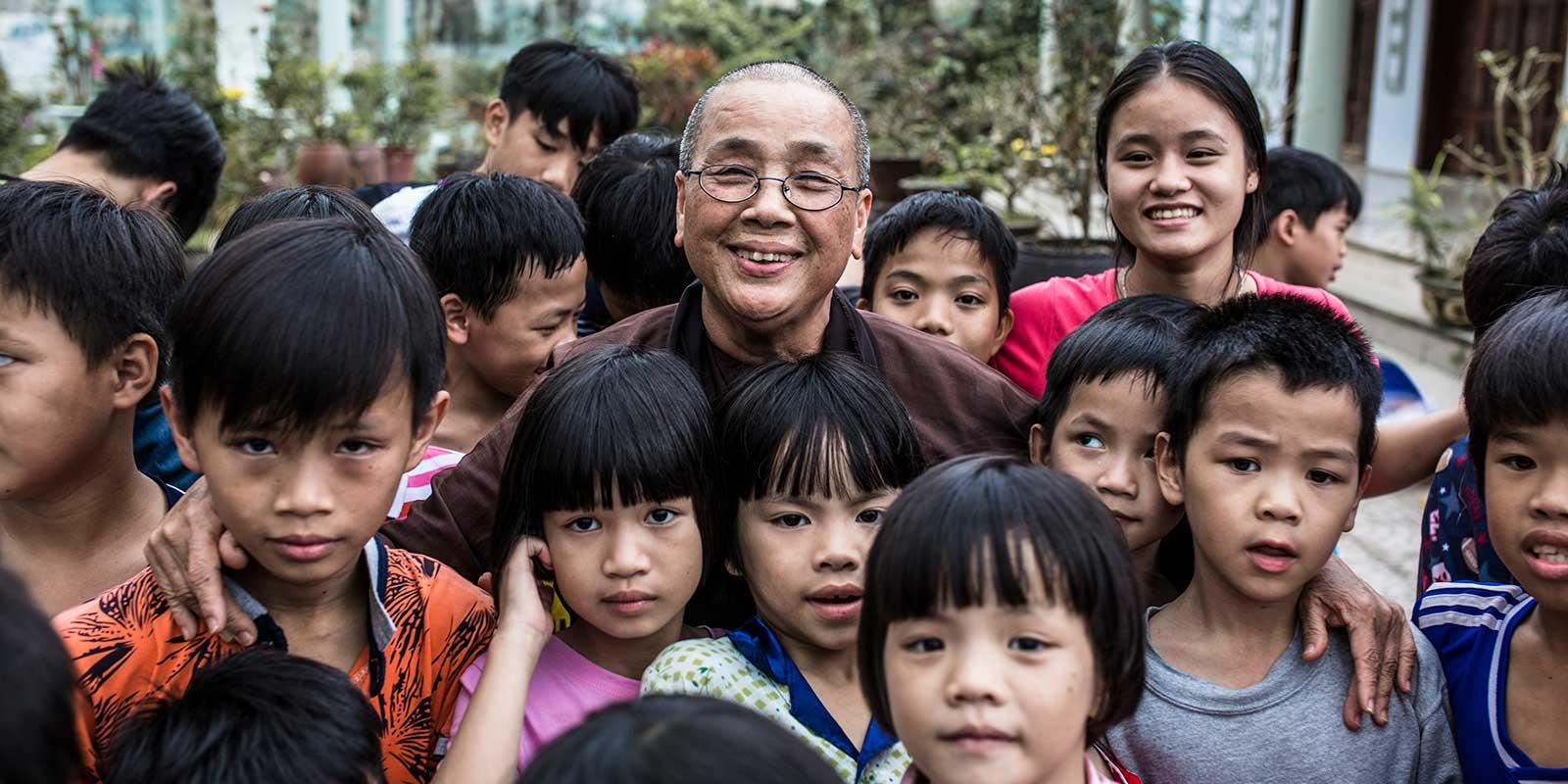A buddhist nun in brown dress and glasses with grouop of children.