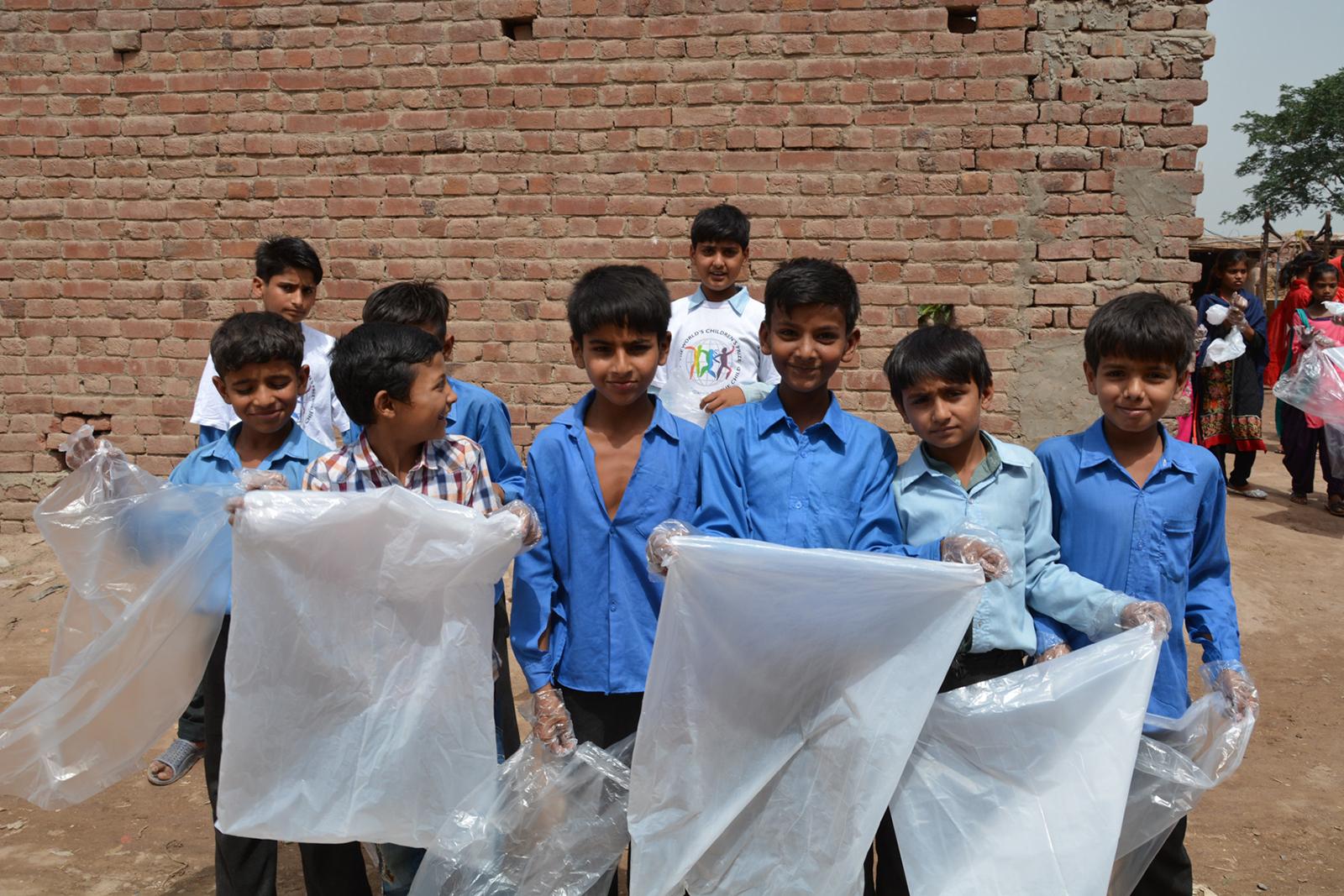 Nine boys in white and blue shirts standing in front of a brick wall, holding plastic bags 