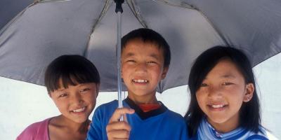Three children under umbrella, smiling. 