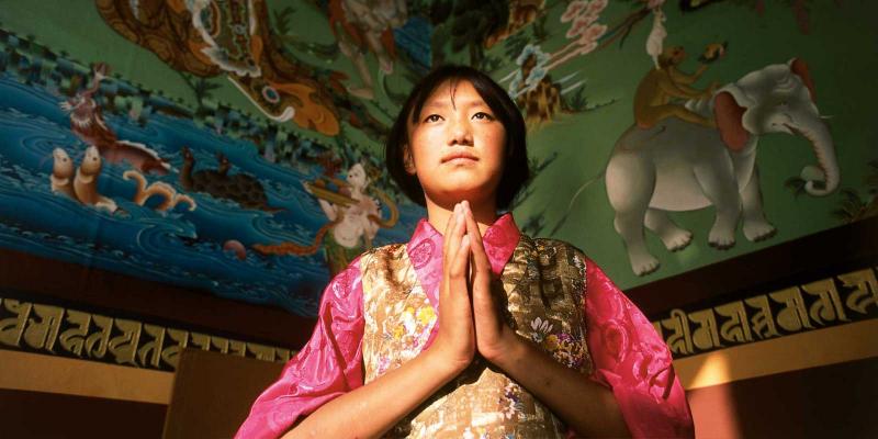 Girl with hands in Buddhist prayer with colorful paintings above her.
