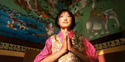 Girl with hands in Buddhist prayer with colorful paintings above her.
