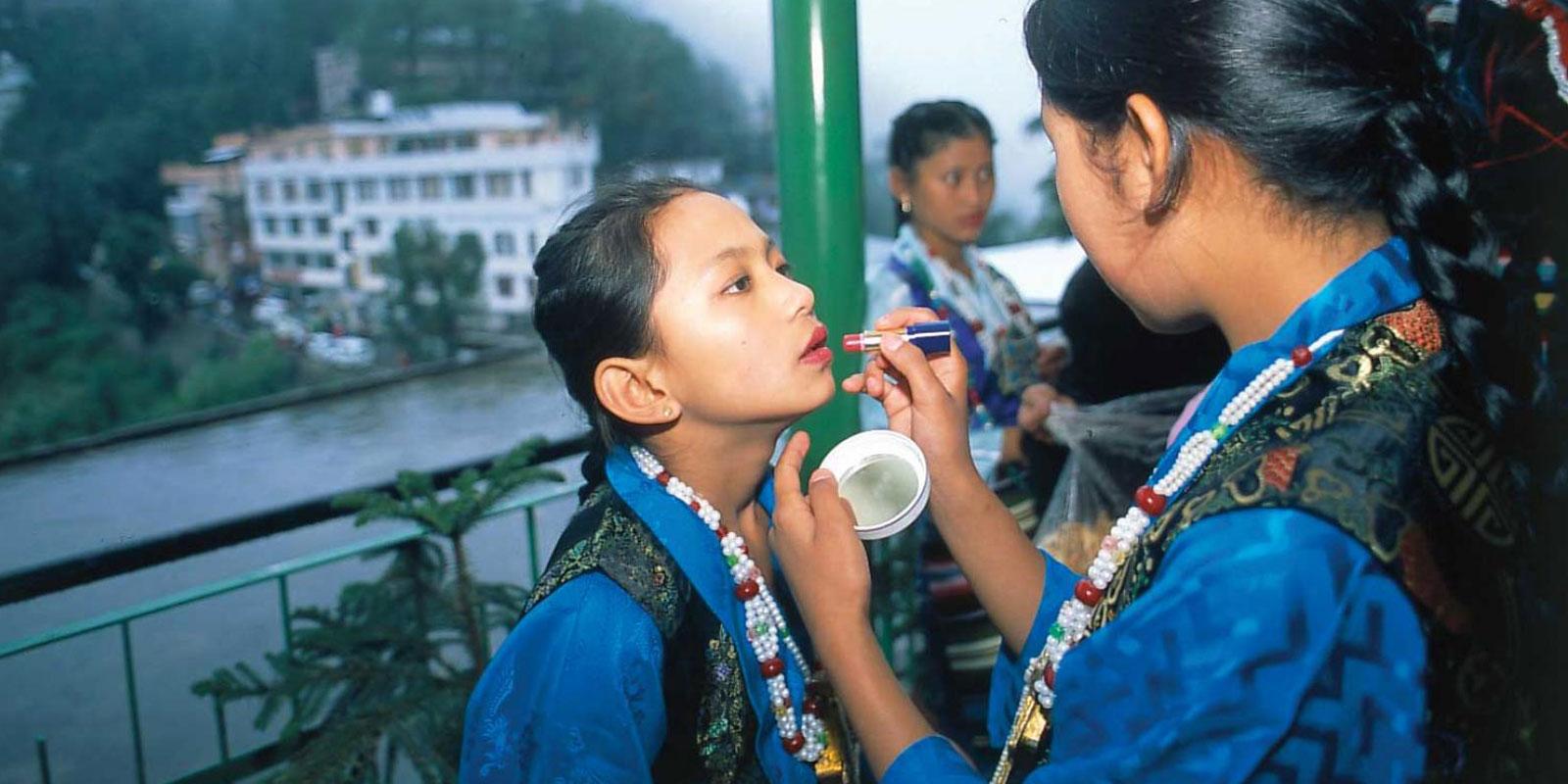 Girl getting her lipstick done by another taller girl, both wearing blue chupa robes and have their hair in a slicked back braid.
